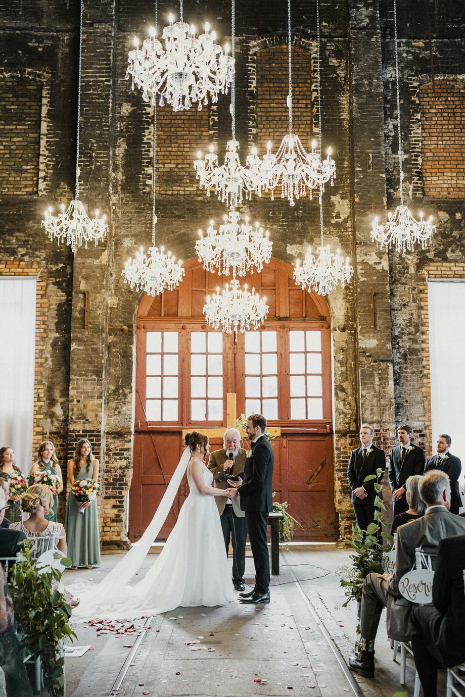 A couple exchanging vows beneath hanging chandeliers in a brick hall
